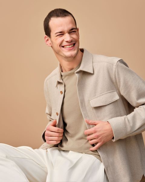 Un homme souriant est assis devant un fond beige, portant une veste marron clair sur une chemise marron clair et un pantalon blanc, et regarde sur le côté avec une expression joyeuse.