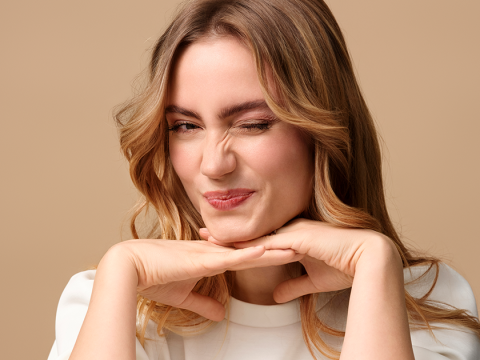 Une personne aux longs cheveux ondulés et au maquillage léger sourit les yeux fermés, le menton appuyé sur les mains. Elle porte un haut blanc devant un fond beige uni.