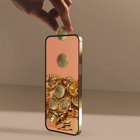 Une main dépose une pièce d'argent dans un smartphone. Sur l'écran, des pièces d'or s'empilent. L'arrière-plan est beige neutre. L'image suggère l'épargne numérique ou l'investissement.