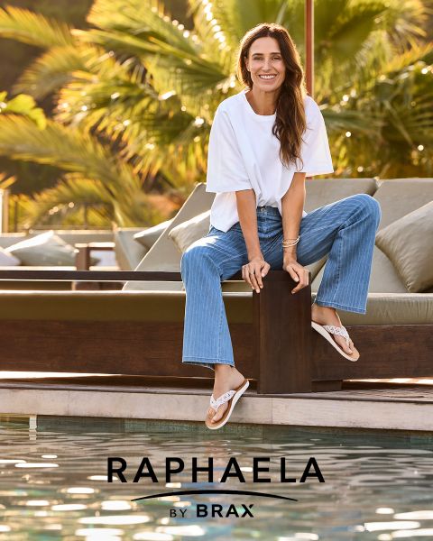 Une femme aux longs cheveux blonds, portant une chemise grise à manches longues et un jean foncé, se tient souriante près d'une balustrade avec un fond bleu clair. À côté d'elle apparaît le texte "EUREX by BRAX".