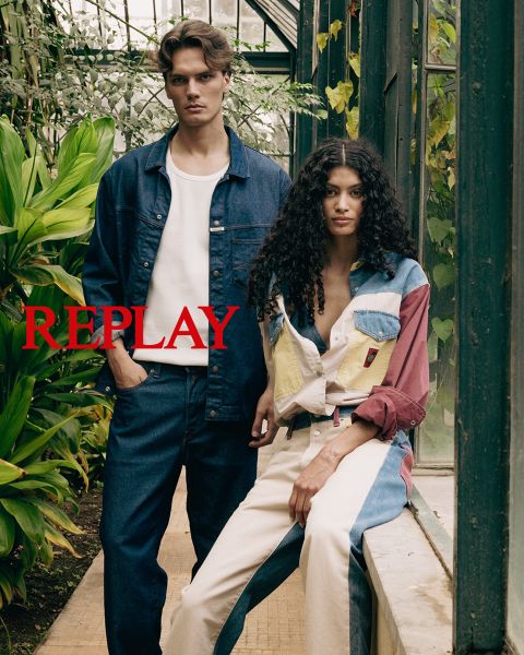 Un homme et une femme, vêtus de vêtements sombres et élégants, sont assis sur un escalier en bois dans un intérieur. Le mot "REPLAY" est affiché bien en évidence au centre de l'image en lettres capitales rouges.