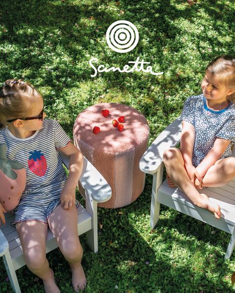 Quatre enfants souriants se tiennent dans un pré et tiennent des bâtons entre lesquels sont tendues des bandes de tissu colorées. Deux alpagas sont à proximité, et à l'arrière-plan, on peut voir un ciel nuageux et des arbres. En haut, il est écrit "Sanetta".