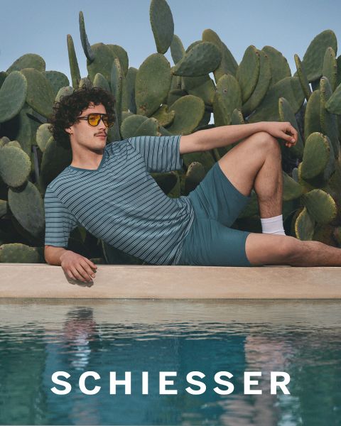 Une femme et un homme en vêtements d'intérieur gris clair sont assis côte à côte sur un tapis et sourient doucement à la caméra. Le nom de la marque "SCHIESSER" est visible dans la partie inférieure de l'image.