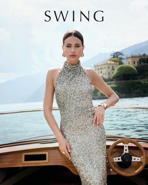 Une femme vêtue d'une robe de soie vert sauge est assise sur une chaise couleur crème devant un fond floral. Elle porte des talons dorés, des boucles d'oreilles voyantes et un bracelet. Le mot "SWING" apparaît en bas de l'image.