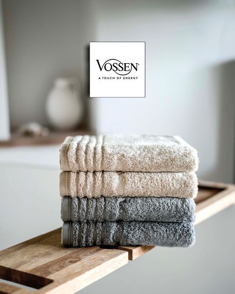 Une femme aux cheveux blonds courts, enveloppée dans une serviette grise, est assise sur un canapé clair et regarde la caméra. Le logo Vossen est visible dans le coin inférieur gauche.