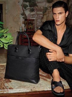 Une personne aux cheveux foncés est assise sur une surface en marbre, portant une chemise boutonnée foncée et un pantalon. Elle est pieds nus, un pied croisé sur l'autre, et porte des sandales noires. Un grand sac en cuir noir est posé à côté d'elle. En arrière-plan, on aperçoit un mur et des meubles de style vintage.