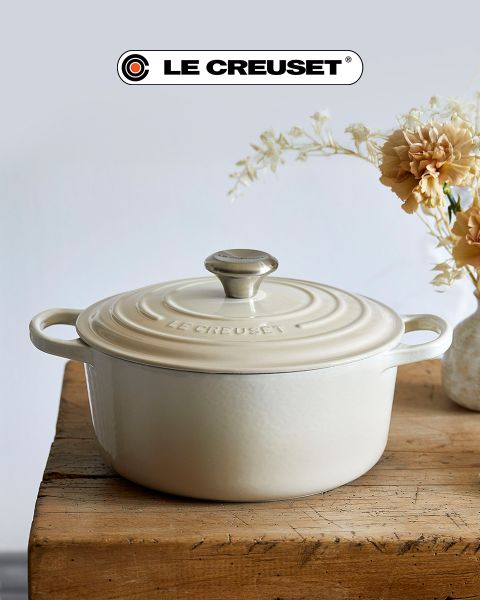 Une scène de cuisine avec une cocotte rouge, une poêle à frire antiadhésive, un ensemble de casseroles empilables en acier inoxydable, une marmite noire et un bol de tomates cerises sur un comptoir en bois clair devant un dosseret en carreaux blancs. Le logo Le Creuset est visible.