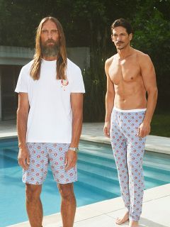 Un homme torse nu, portant un blazer noir et un short foncé, est assis sur une chaise blanche au bord de falaises rocheuses face à la mer, tenant un verre de vin blanc à la main, tandis que les vagues se brisent en arrière-plan.