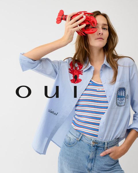 Une femme portant un pull rouge avec un cœur blanc, un jean bleu et un bonnet blanc pose devant un mur clair. En bas de l'image, le mot « oui » est écrit en lettres blanches.