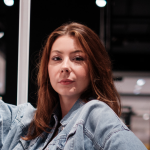 Une personne aux longs cheveux bruns regarde la caméra. Elle porte une veste en jean. L'arrière-plan est flou, ce qui suggère une prise de vue en intérieur.