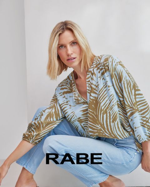 Une femme pose devant un mur blanc, portant un gilet rouge sans manches sur un chemisier rayé rouge et blanc et un pantalon foncé. En bas de l'image, on peut lire le mot "RABE".