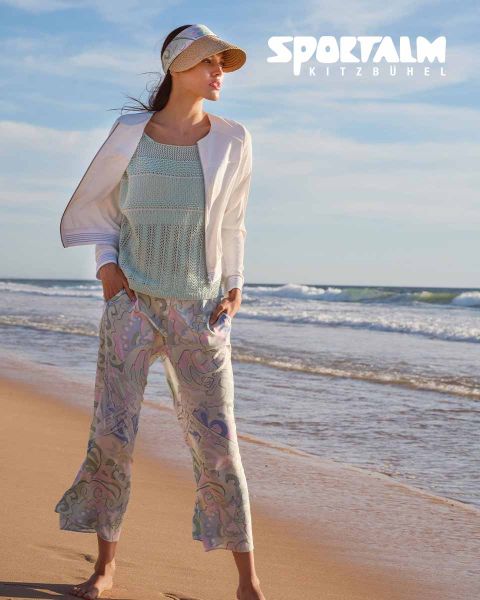 Une femme se tient sur une plage. Elle porte un haut en tricot bleu clair, une veste blanche et un pantalon à motifs. Elle porte une visière et regarde vers la mer. Dans le coin supérieur droit, on peut lire « SPORTALM KITZBÜHEL ».