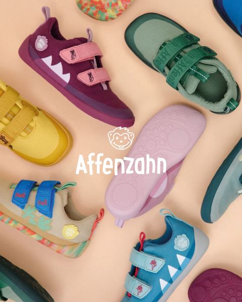 Un enfant portant un pull orange vif et un pantalon rayé est assis sur une surface colorée et pointe un avion en papier de style bande dessinée dans le ciel. Il sourit et est entouré de nuages duveteux. Dans le coin, on peut lire « Affenzahn ».