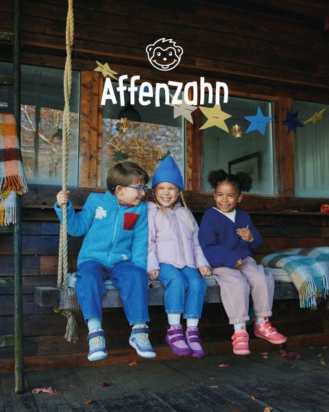Un enfant portant un pull orange vif et un pantalon rayé est assis sur une surface colorée et pointe un avion en papier de style bande dessinée dans le ciel. Il sourit et est entouré de nuages duveteux. Dans le coin, on peut lire « Affenzahn ».