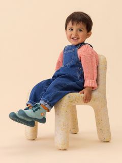Un jeune enfant en chemise rayée et salopette en jean est assis avec une jambe à l’extérieur et montre la semelle bleue texturée de sa chaussure.