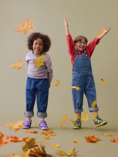 Un pied d’enfant nu à côté d’une chaussure de sport avec des bandes jaunes, tous deux visibles sous un pantalon à carreaux vert clair sur un sol beige.