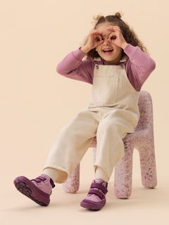 Un enfant souriant avec un bonnet vert, une chemise rayée et un gilet est assis sur un bloc couleur pêche devant un fond jaune, portant des bottes et un pantalon clair.