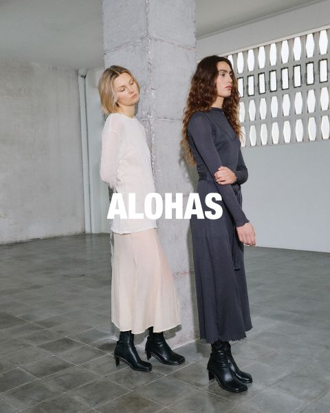 Une femme portant un haut court blanc et un pantalon beige avec des accents orange est assise sur un tabouret. Elle se trouve dans une pièce rustique avec de grandes fenêtres. Au centre, on peut lire le texte "ALOHAS".