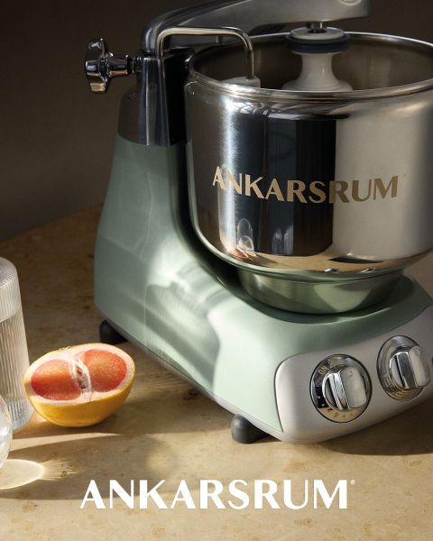 Un robot culinaire Ankarsrum brillant avec un bol en acier inoxydable et une base rose est présenté sur une surface rose assortie, avec le nom de la marque « ANKARSRUM » visible à la fois sur le bol et sur le fond.