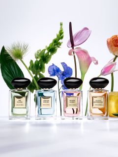 Quatre flacons de parfum sont exposés sur et autour d'un socle beige. Les flacons sont carrés avec des bouchons arrondis et sont de couleur noire et rouge. Un mur blanc ondulé et texturé sert de fond.