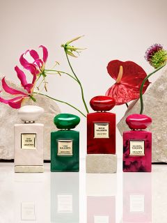 Huit flacons de parfum de différentes couleurs, dont rouge, vert, bleu, rose et noir, sont disposés sur des socles beiges devant un fond clair. Chaque flacon porte une étiquette avec une écriture dorée.