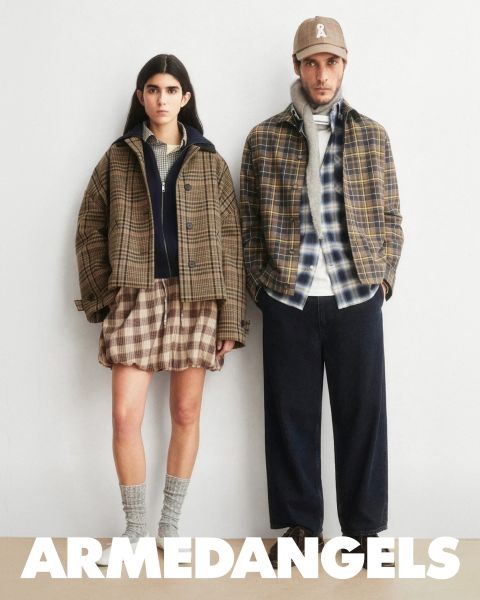 Deux jeunes adultes en vêtements en jean se tiennent ensemble en plein air, l'un avec le bras levé, l'autre avec une valise couverte d'autocollants. En bas, en lettres blanches grasses, il est écrit "ARMEDANGELS".