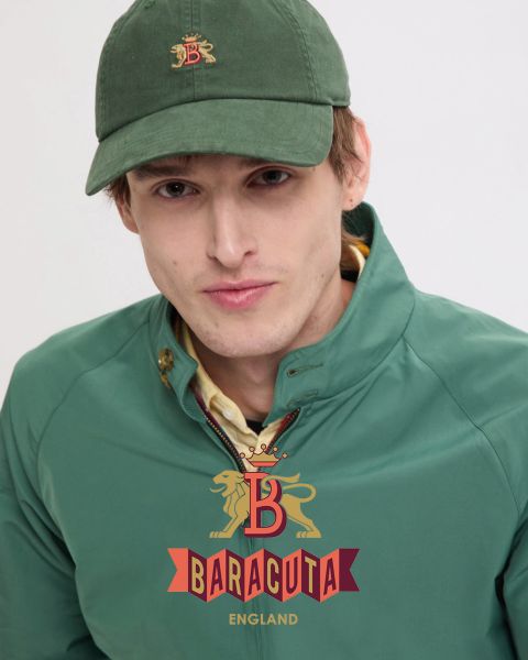 Une personne aux cheveux courts et foncés, portant une boucle d'oreille et une veste foncée sur une chemise blanche, regarde la caméra. L'intérieur de la veste est à carreaux rouges. Le logo Baracuta avec un lion et un "B" est visible sur le devant.