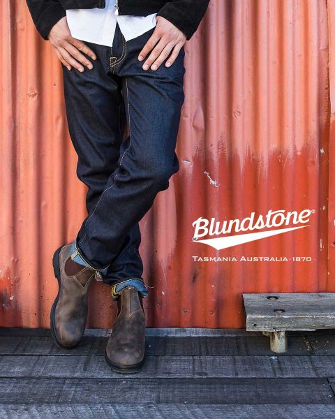 Une personne portant des bottes Blundstone marron usées se tient sur un bateau délabré près de cordages, avec de l'eau et des rochers flous en arrière-plan.