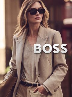 Une femme aux longs cheveux bruns et au maquillage neutre porte une veste en peau de mouton retournée marron et un pull à col roulé beige, et regarde directement la caméra avec une expression calme, devant un fond uni.