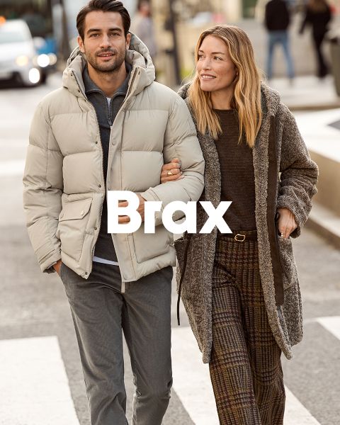 Une femme portant un manteau à motifs et un pull rouge se tient à côté d'un homme portant un manteau d'hiver bleu marine et un pantalon beige. Ils sont appuyés contre un mur de pierre à l'extérieur. Le mot "Brax" apparaît en bas de l'image.