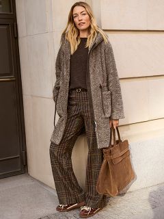 Une femme aux cheveux longs se tient sur un trottoir en ville. Elle porte un pull noir à rayures blanches et un pantalon noir à jambes larges, et tient une grande pochette noire à la main, tandis qu'elle est éclairée par le soleil venant de derrière.