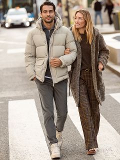 Une femme portant un manteau de fourrure à motifs et un haut rouge se tient souriante à côté d'un homme portant une veste d'hiver bleu marine et un pull beige. Ils se tiennent à l'extérieur, près d'un mur de pierre, et la femme tient un sac à main marron.
