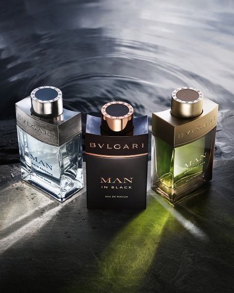 Une femme en robe blanche se penche en arrière devant un décor architectural moderne sous un ciel bleu. Au premier plan, deux flacons du parfum Bvlgari Omnia Crystalline, aux teintes claires et ambrées.