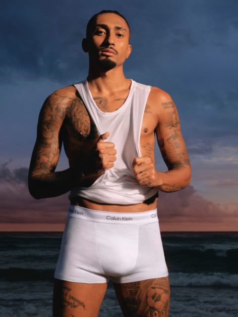 Un homme en veste noire et jean se tient dans un stade, les mains dans les poches, et regarde sur le côté. On aperçoit des sièges de stade et un grand bâtiment en arrière-plan.