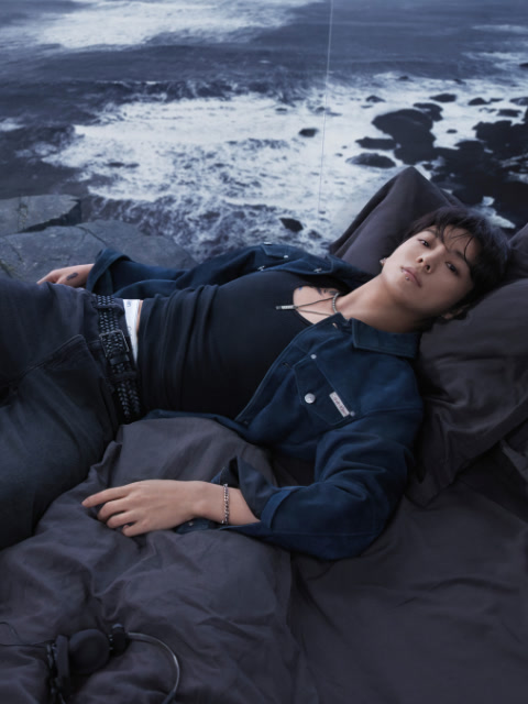 Photo en noir et blanc d'une femme portant un soutien-gorge de sport Calvin Klein et un jean, allongée sur le dos, les jambes pliées au-dessus de la tête, les bras tendus sur le sol, le regard fixé sur l'appareil photo.