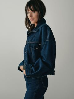 Une personne aux longs cheveux blonds est assise sur un banc en bois, portant une veste en jean bleue, une chemise blanche et un jean ample. En arrière-plan, on aperçoit des bâtiments urbains et des gratte-ciel.