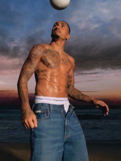 Un homme portant une veste en jean à motifs et un jean bleu se tient sur un pont avec des balustrades métalliques. En arrière-plan, on aperçoit une rivière et un autre pont par temps nuageux.