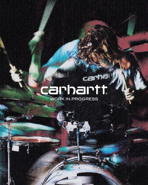 Image en noir et blanc d'une personne portant un chapeau et une veste, accroupie avec les mains sur les genoux. Le texte "Carhartt Work In Progress" est clairement visible au centre.