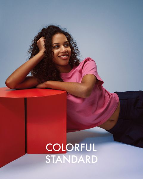 Une personne aux longs cheveux blonds se tient de profil devant un fond bleu. Elle porte un t-shirt bleu foncé, un jean bleu clair et des baskets. Le texte « COLORFUL STANDARD » est affiché en blanc.