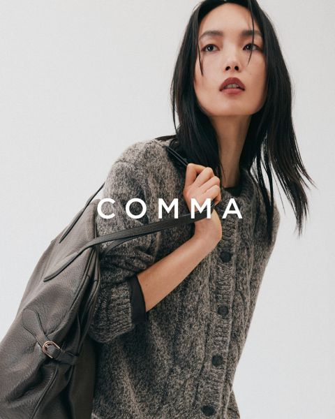 Une femme portant un manteau à carreaux et des accessoires dorés pose avec un chien beige et blanc portant une tenue à carreaux assortie. Le mot « COMMA » apparaît en grandes lettres blanches sur l'image.
