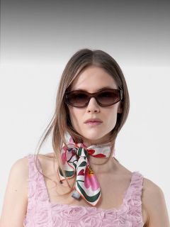 Femme portant une robe rose texturée avec des motifs de rosettes, avec des lunettes de soleil foncées et un foulard coloré noué autour du cou, debout devant un fond uni.