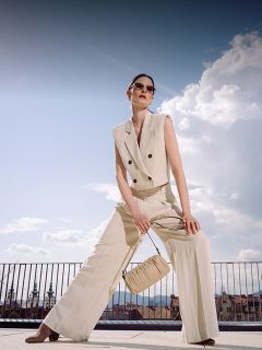 Une femme portant un tailleur pantalon beige sans manches et des lunettes de soleil pose en extérieur, tenant un sac assorti, avec un paysage urbain et un ciel bleu en arrière-plan.