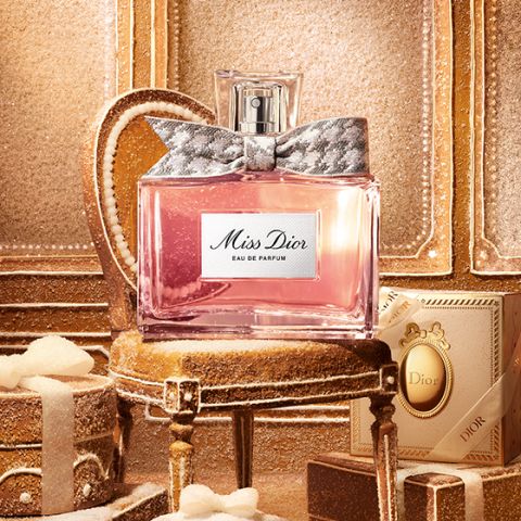 Un flacon en verre transparent de « Miss Dior Eau de Parfum », rempli d'un liquide rose et orné d'un nœud argenté à motifs autour du col du flacon, sur un fond blanc uni.