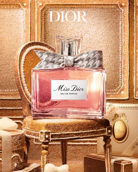 Un homme tatoué et orné de bijoux est appuyé sur une table à côté d'une bouteille de parfum « Dior Sauvage Extrait » devant un décor extérieur accidenté.