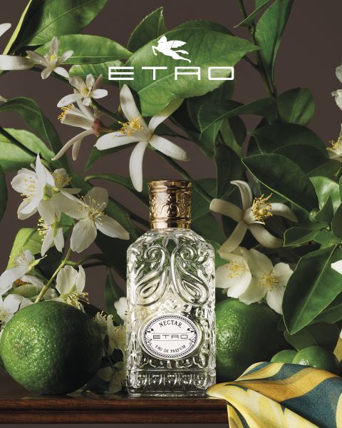 Flacon de parfum portant l'inscription "ETRO" et un motif de pomme de pin, entouré d'un décor artistique : une sphère, un œuf avec un papillon, une pierre, une feuille, des noix, une pomme dorée et une main sculptée. En haut, le logo ETRO devant un fond de ciel bleu et de mer.