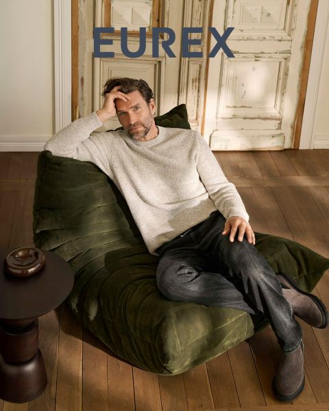 Un homme se tient à l'extérieur près de hautes herbes, portant un pull beige, une veste sans manches bleu marine et un pantalon marron, et regarde sur le côté. Le logo "EUREX by BRAX" apparaît sur le côté droit. Le ciel est nuageux.