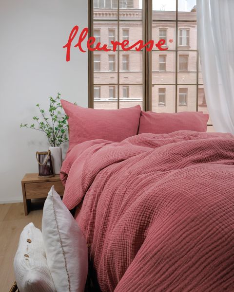 Une chambre confortable avec un lit bien fait, recouvert de draps blancs à motifs d'oiseaux. Deux coussins décoratifs sont posés sur la tête de lit. La lumière du soleil filtre à travers des rideaux transparents, projetant une douce lueur sur le plancher en bois clair.