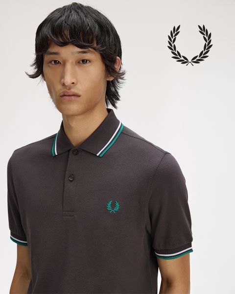 Une personne aux longs cheveux tressés regarde pensivement vers le haut tout en ajustant une chemise blanche à col. La chemise est ornée d'une couronne de laurier. L'image est en noir et blanc.