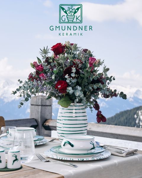 Une table magnifiquement dressée avec des assiettes en céramique décoratives ornées de motifs abstraits de lignes noires. Des feuilles d'eucalyptus sont disposées sur les assiettes. Une fourchette dorée, un pot à crème rayé et une tasse à motifs complètent l'élégante nappe rose.