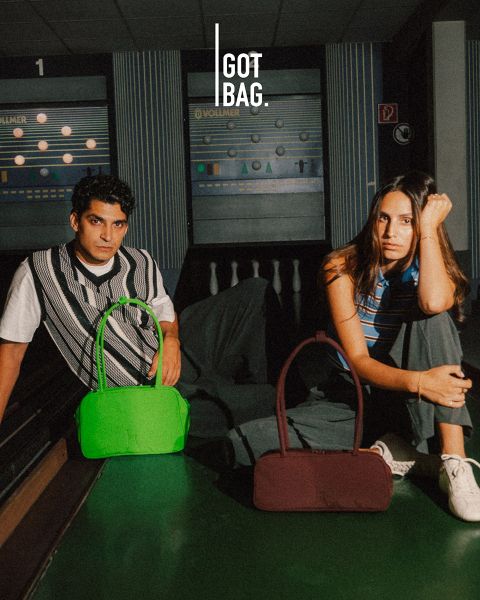Un homme portant des lunettes de soleil tient de grands sacs gris devant un fond à rayures foncées. En dessous de lui, différents sacs de différentes tailles et couleurs sont soigneusement exposés. Le logo en haut à droite est « GOT BAG ».